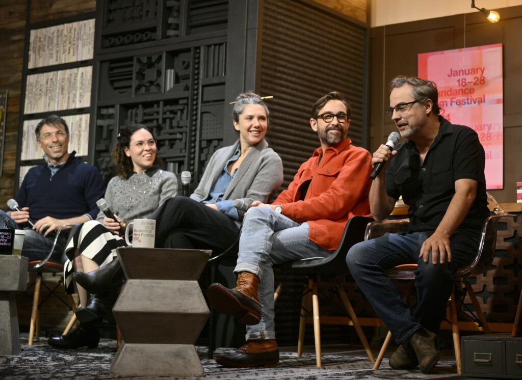 Dr. Martin Monti, Dr. Monica Lopez, Sam Zuchero, and Andy Zuchero look smiling to their right as Alex Rivera speaks into a microphone. They are all sitting down.